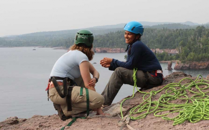 Two people wearing safety gear are secured by ropes near the edge of a cliff. One person appears to be an instructor, giving direction to the other person. There is a lake below them.